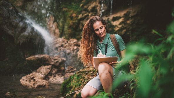 photo female biologist kneels in nature to conduct an ecological survey on a clipboard