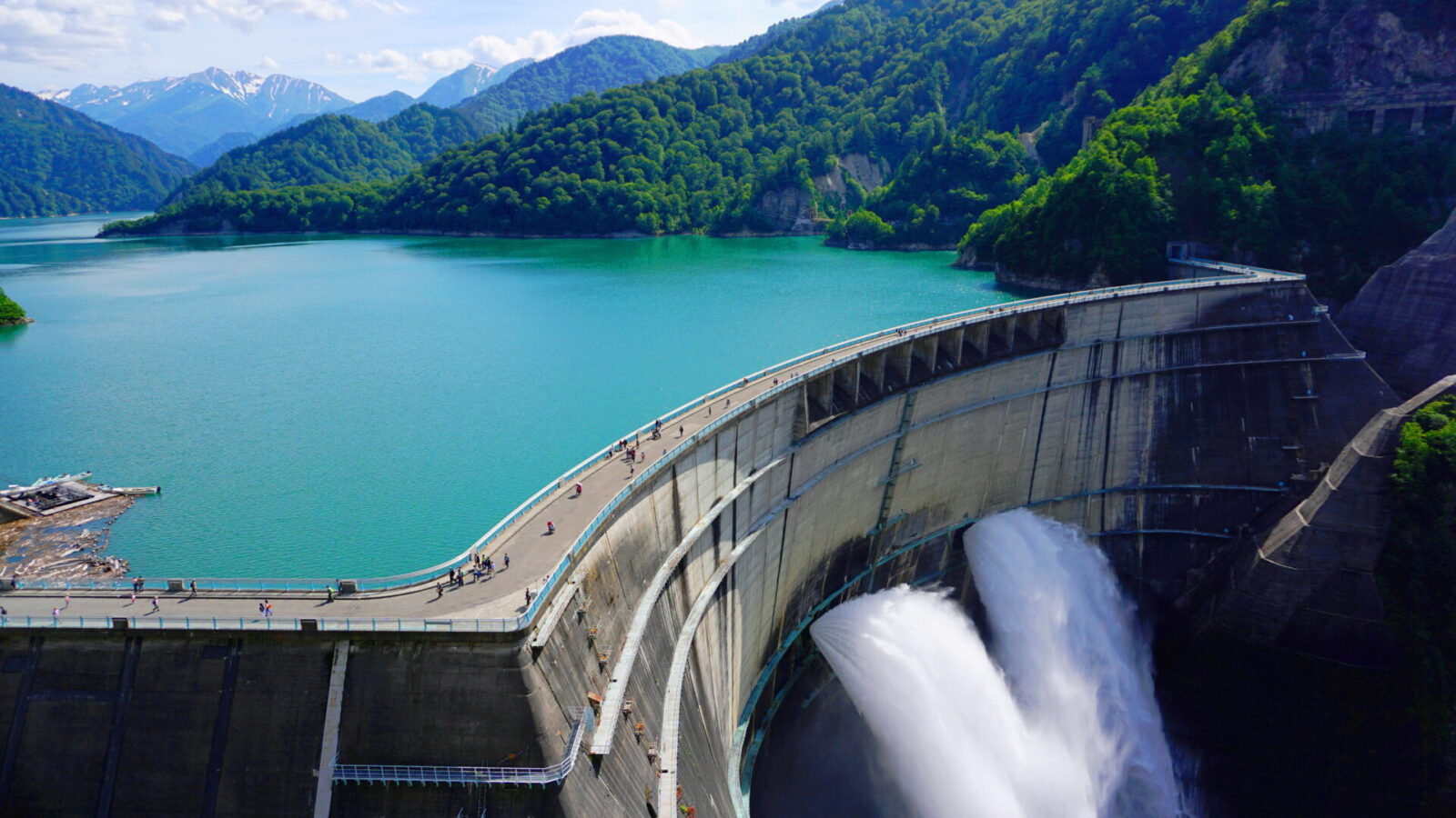 photo of lake / reservoir surrounded by green alpine mountains; Kurobe Dam flood gates are open