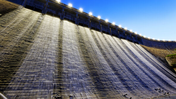 photo hydropower generation station at dusk | camera angle looks up a dam and a line of lights along the top of the dam