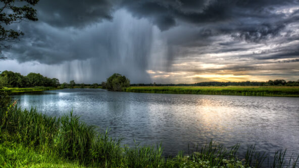 photo dark clouds and intense rainstorm in the distance; relatively calm river and vibrant green grasses in the foreground