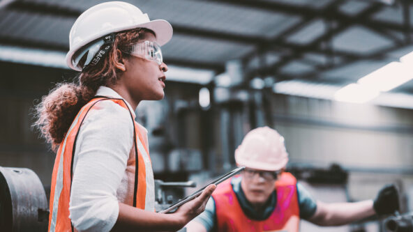 photo female engineer wears a white hard hat and reflective orange safety vest, holds a tablet while working in a water facility, a water operator works in the background