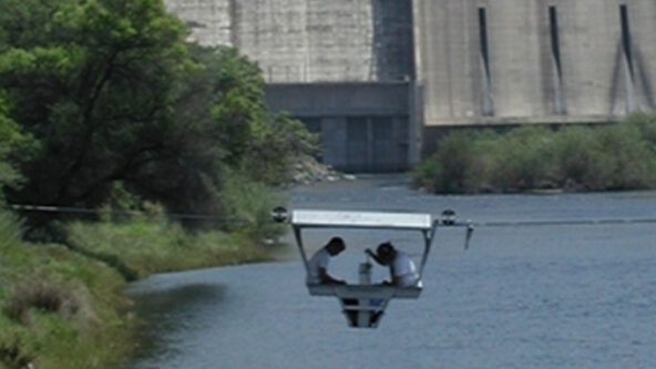 photo of two hydrographers in a gondola taking discharge measurements with a hydropower dam in the background | source Sierra Hydrographics