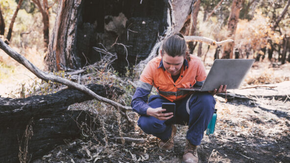 photo of a female field inspector holding a mobile phone and a laptop in addition to examining the ground and soil